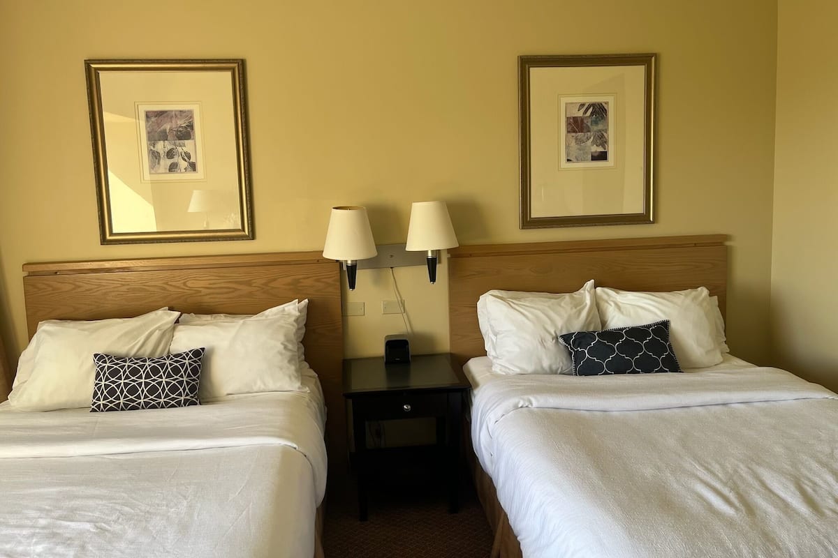 A hotel room with two beds, each with white linens and decorative pillows. There are framed pictures on the wall above each bed.