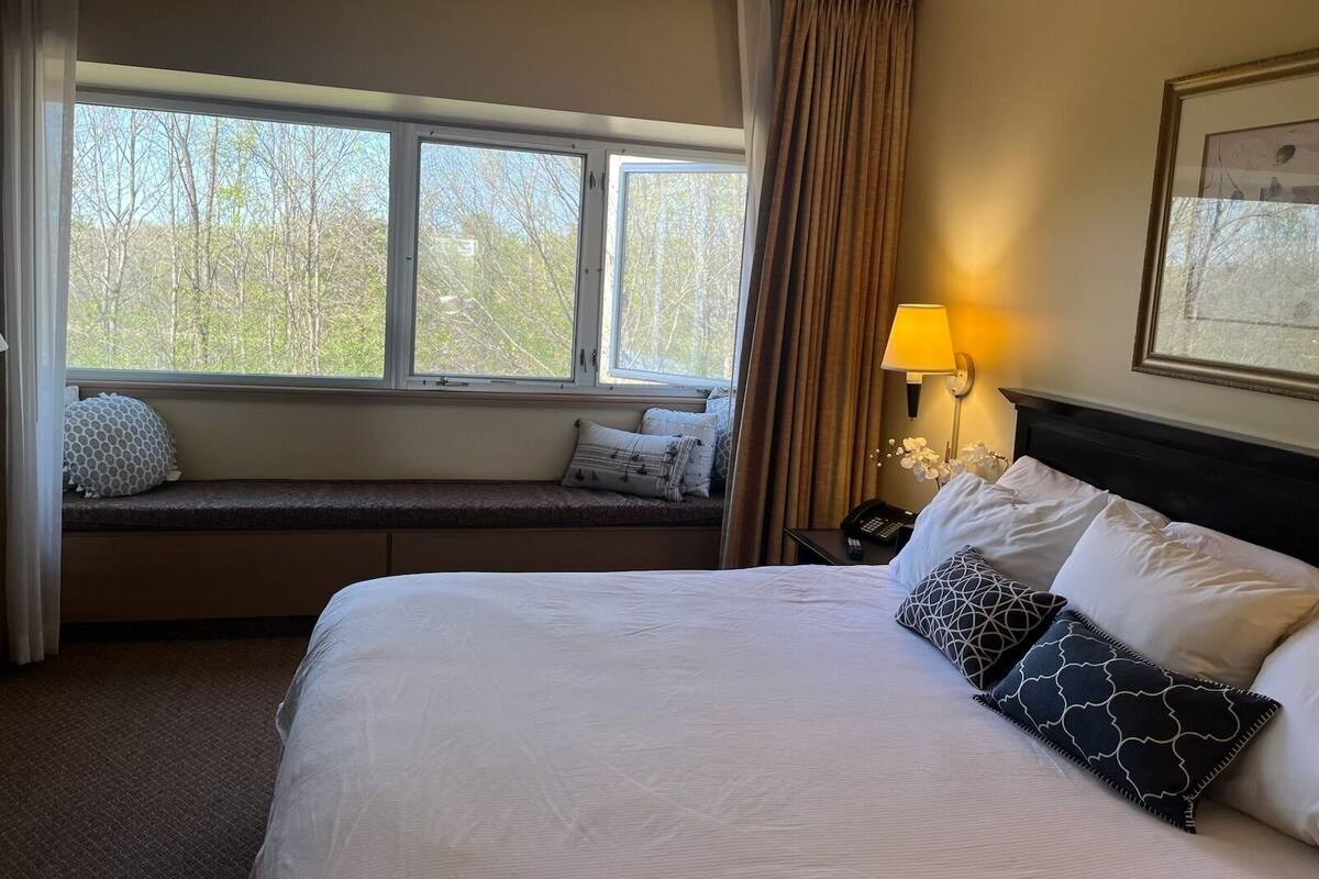 A hotel room with a large bed, a window seat with pillows, a bedside table with a lamp, and a framed picture on the wall.