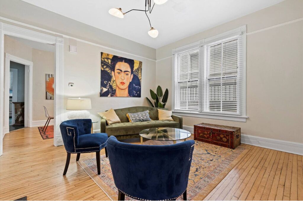A living room with a green sofa, two blue armchairs, a glass coffee table, a painting on the wall, and a window with blinds.