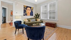 A living room with a green sofa, two blue armchairs, a glass coffee table, a painting on the wall, and a window with blinds. - 4 Bed 1.5 Bath, 3 Story Townhouse, Free Parking (Milwaukee)