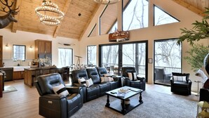 A spacious living area with leather sofas, a coffee table, and a chandelier. Large windows provide natural light and views of the outdoors. - Modern Cabin, Cozy and Peaceful (Lake Geneva)