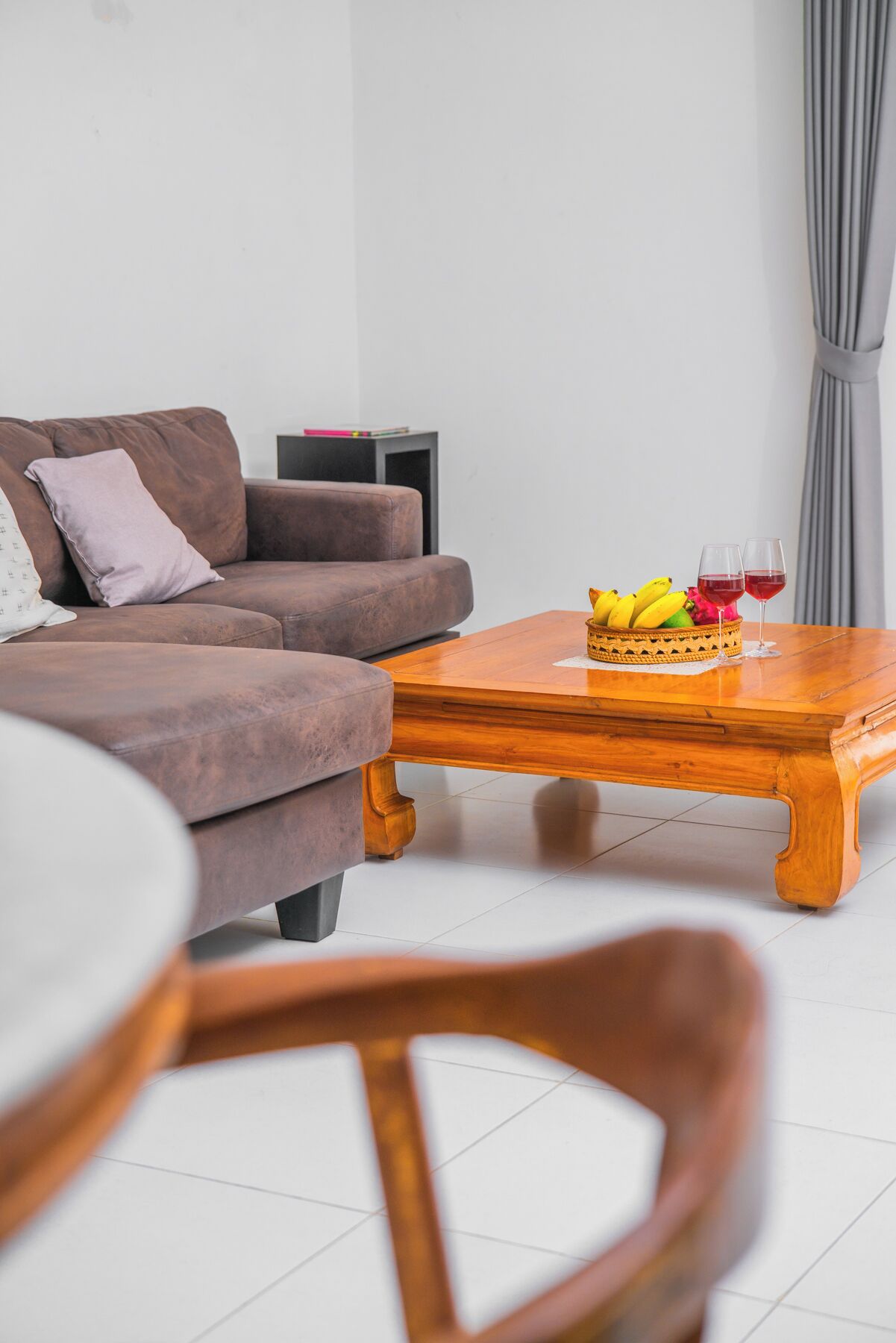 A living room with a brown sofa, a wooden coffee table with fruit and wine glasses, and a dining table with chairs.