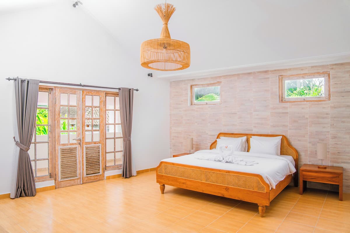 A bedroom with a wooden bed, a hanging light fixture, and two windows with views of greenery.