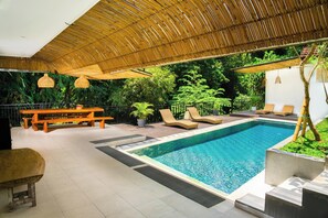 Un espace piscine aménagé avec des chaises longues, un banc en bois et une structure dotée d’un toit de chaume.