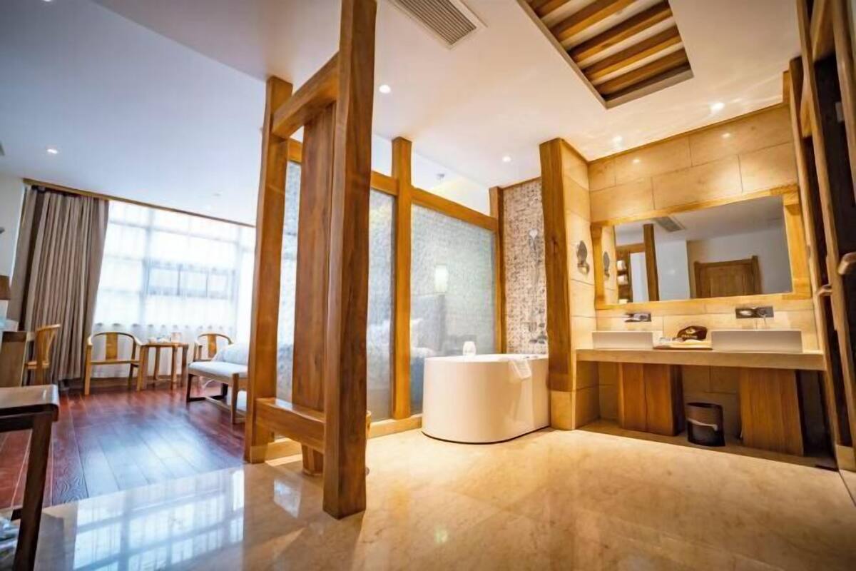 A modern hotel room with a wooden floor, a large window, a sitting area with chairs, a bathtub, and a vanity with a mirror.