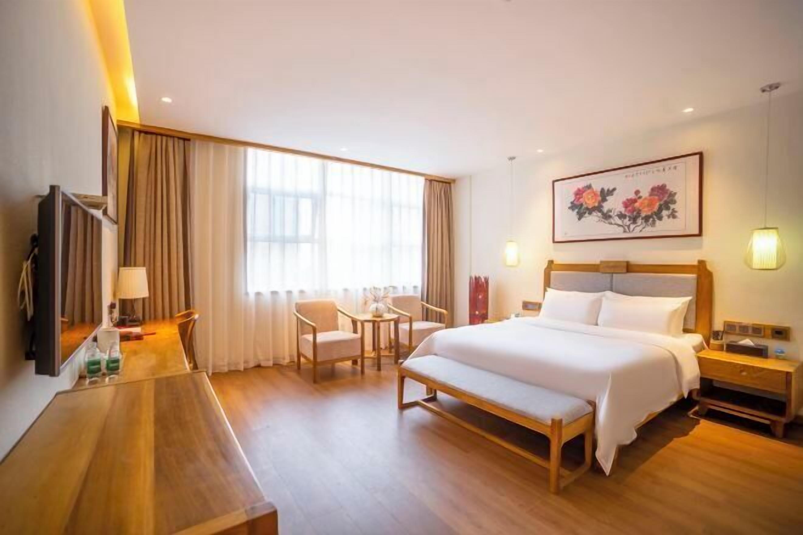 A hotel room with a bed, bedside tables, a desk, chairs, a TV, and a large window with curtains.