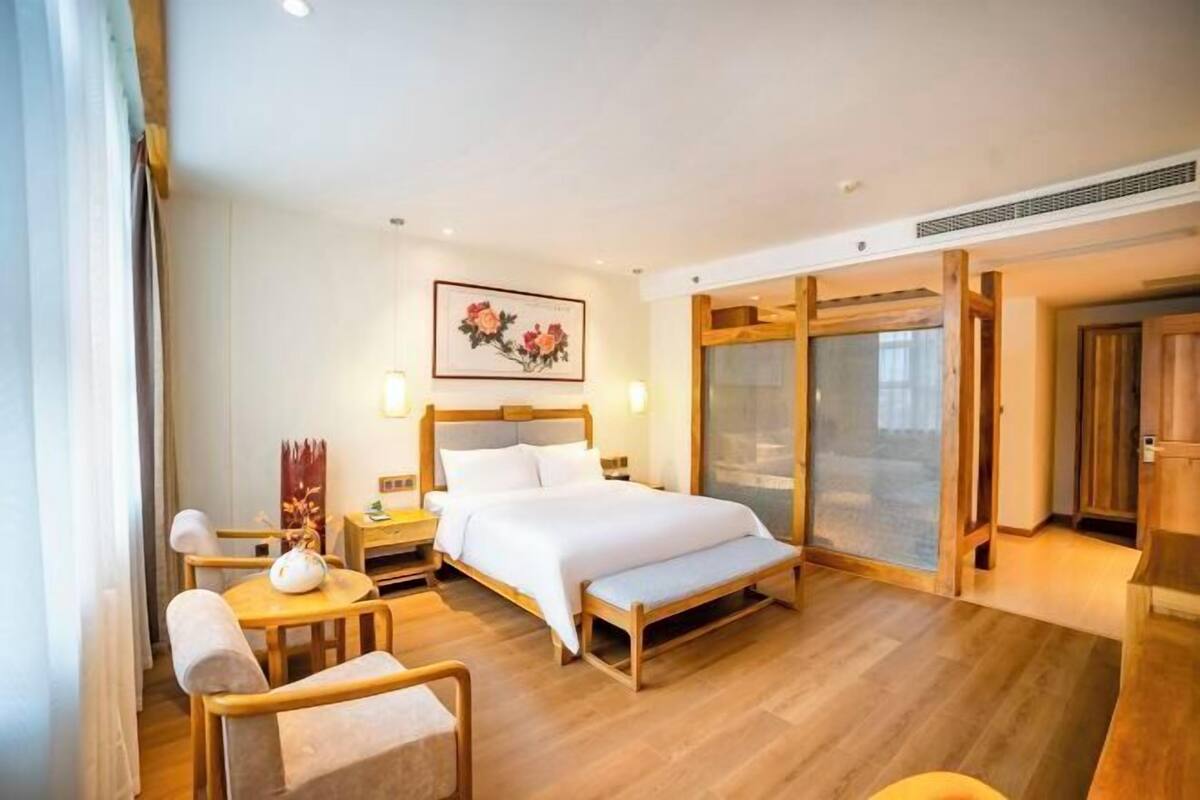 A hotel room with a bed, a bedside table, a chair, a bench, a painting on the wall, and a sliding door.