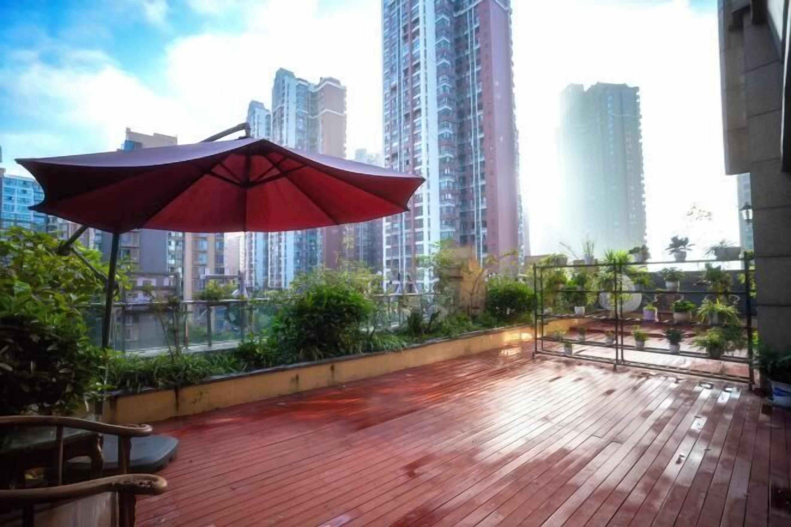 A wooden deck with a red umbrella, surrounded by greenery and tall buildings in the background.
