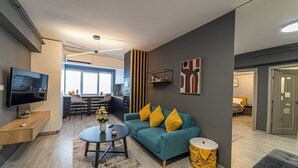 Ein modernes Wohnzimmer mit einer Couch, Couchtisch und Fernseher. Im Hintergrund ist eine Küchenzeile mit Esstisch und Stühlen zu sehen.