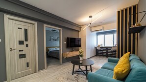 Ein modernes Wohnzimmer mit einem Türkis-farbenen Sofa, einem kleinen Couchtisch und einem Fernseher. Eine Holztür mit Glasfeldern, ein Bett im Hintergrund sowie ein Essbereich mit Tisch und Stühlen sind ebenfalls zu sehen.