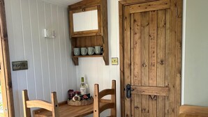 Una mesa de madera con sillas, un estante con tazas y una botella de vino.