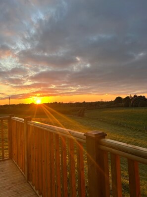 Une terrasse en bois offrant une vue sur un coucher de soleil au-dessus d’un champ verdoyant.