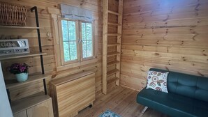 A wooden cabin interior with a green sofa, a floral rug, a window with a view, and wooden shelves with a coffee machine. - Off-grid TWO Glamping Bunkies on Bras d’Or - Marble Mountain/Lime Hill (Lime Hill)