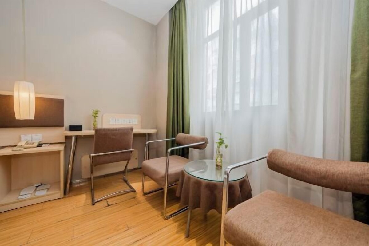 A modern hotel room with a wooden floor, a desk with a lamp, two chairs, a round glass table, and a view through sheer curtains.