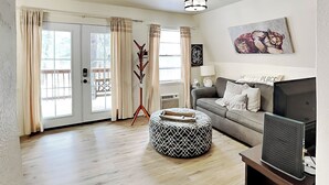 Un salon avec un canapé gris, un pouf rond, un téléviseur posé sur un meuble, un porte-manteau et un tableau accroché au mur.