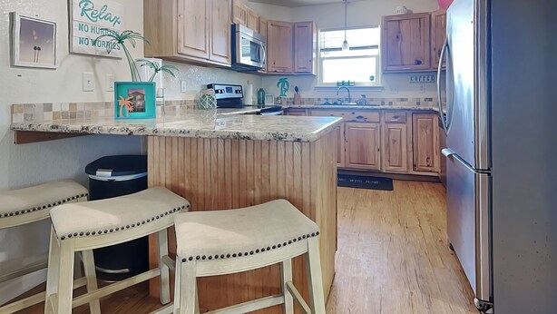 A kitchen with wooden cabinets, a stainless steel refrigerator, a microwave, and a bar stool area. - Twin Palms at Surfside by Ryson Vacation Rentals (Freeport)