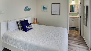 A bedroom with a white bed, a blue pillow with "SURF" text, a wall-mounted light, and a small table with a lamp. - Twin Palms at Surfside by Ryson Vacation Rentals (Freeport)
