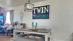 A dining area with a table, chairs, and a bench. There is a wall decoration with the words "TWINS PALMS". - Twin Palms at Surfside by Ryson Vacation Rentals (Freeport)