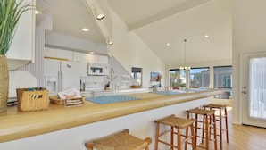 A modern kitchen with a long island counter, bar stools, and a view of the outside. - Beachfront Dolphin by Ryson Vacation Rentals (Galveston)