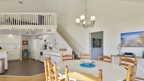 A modern kitchen with white cabinetry, a dining area with a round table and wooden chairs, and a staircase leading to an upper level. - Beachfront Dolphin by Ryson Vacation Rentals (Galveston)