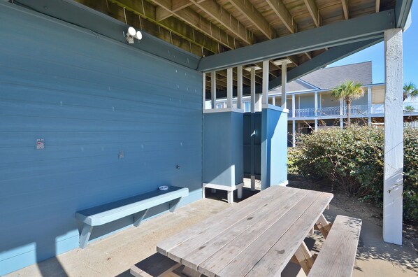 Un espace extérieur abrité avec un banc en bois et une table de pique-nique, un bâtiment bleu en arrière-plan et un palmier.