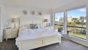 A bedroom with a large bed, a nightstand, a lamp, and a window with a view of palm trees and buildings. - Beachfront Dolphin by Ryson Vacation Rentals (Galveston)