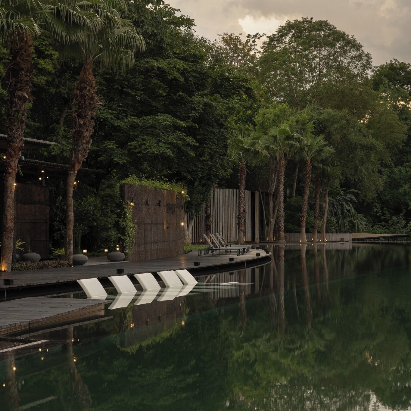 Un espace piscine extérieur paisible, doté d’une terrasse en bois, de palmiers et d’une structure en pierre.