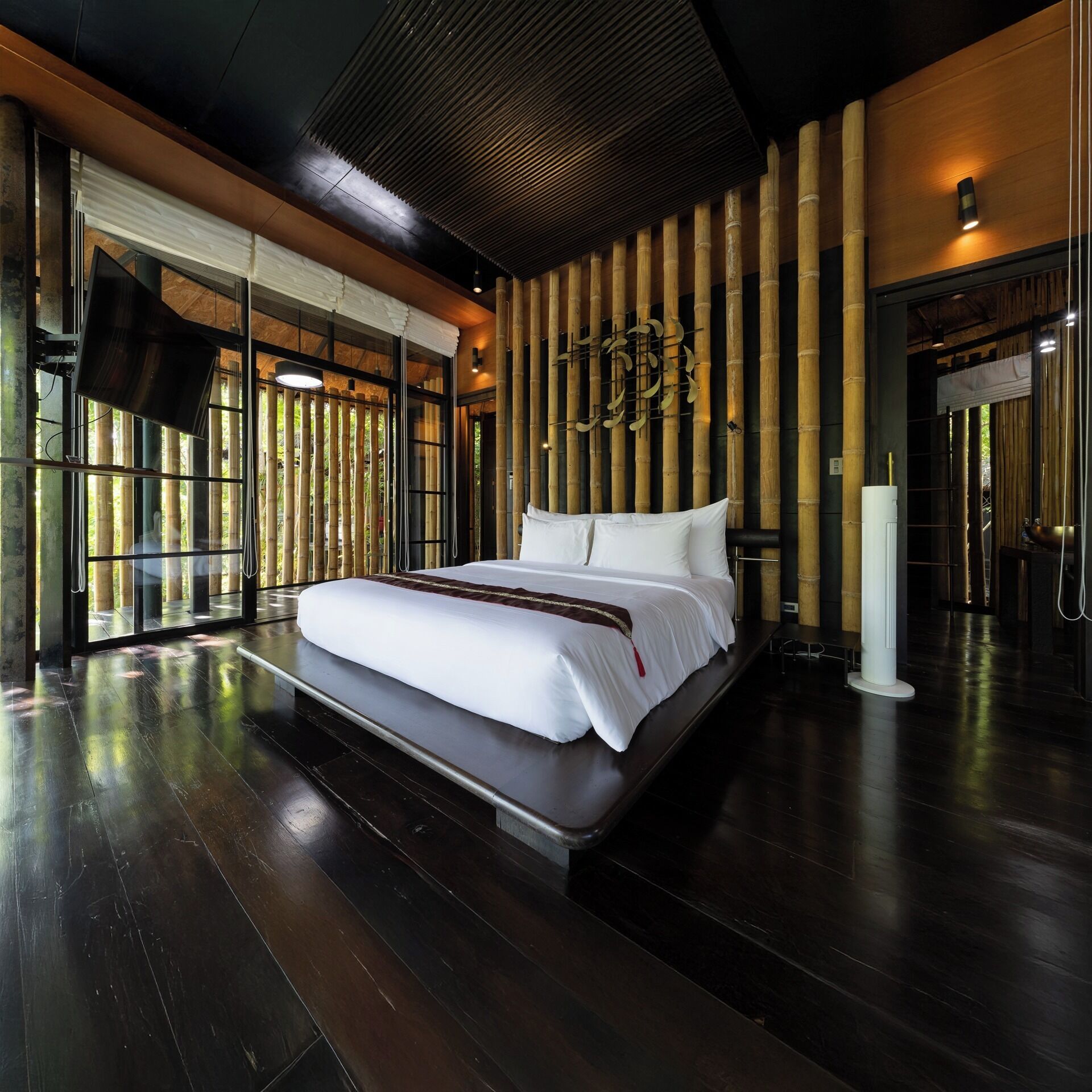 A modern hotel room with a large bed, a flat-screen TV, and a view of bamboo outside the window.