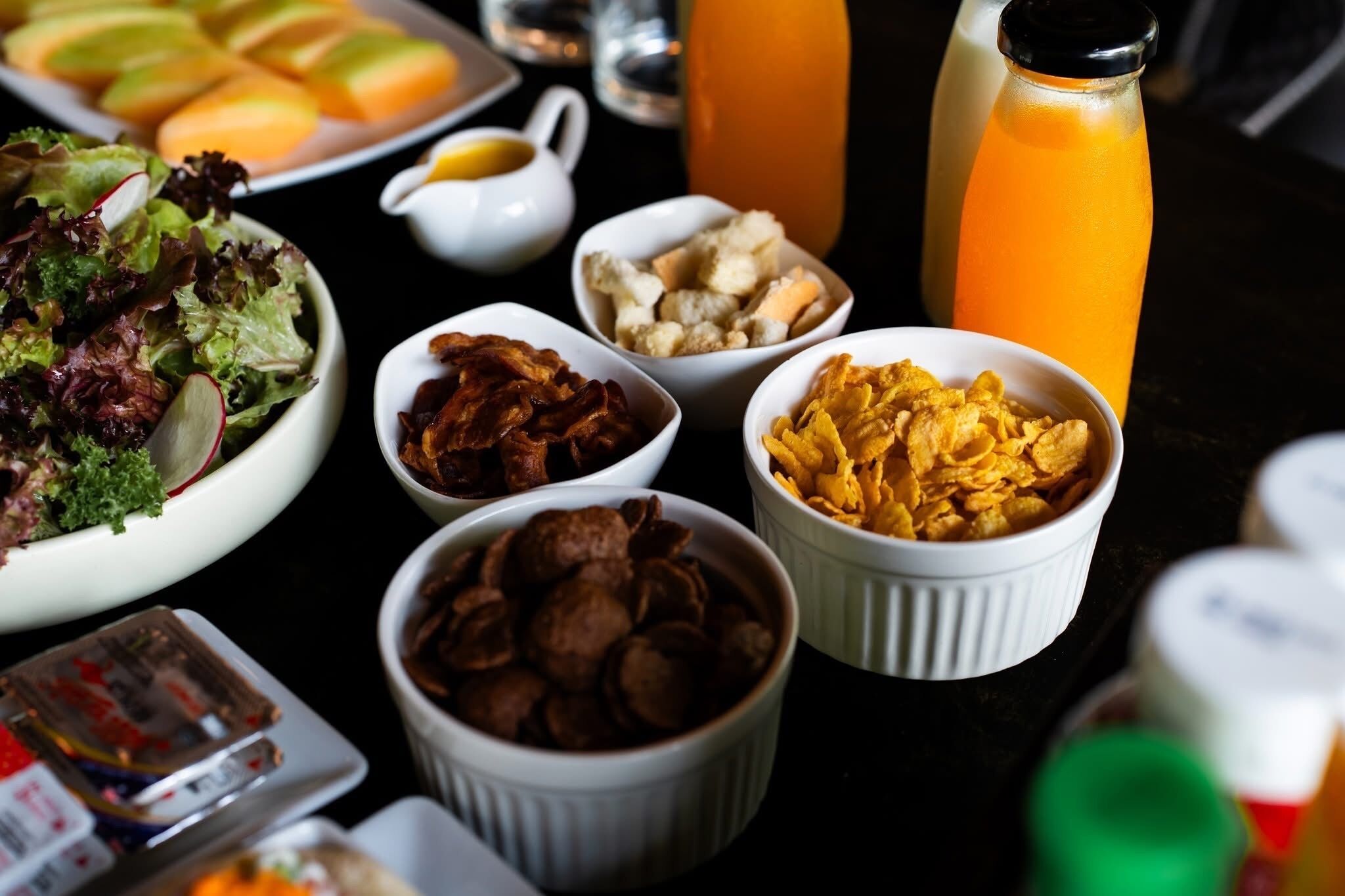 Uma mesa com diversos alimentos, incluindo salada, bacon, cereais e uma garrafa de suco de laranja.