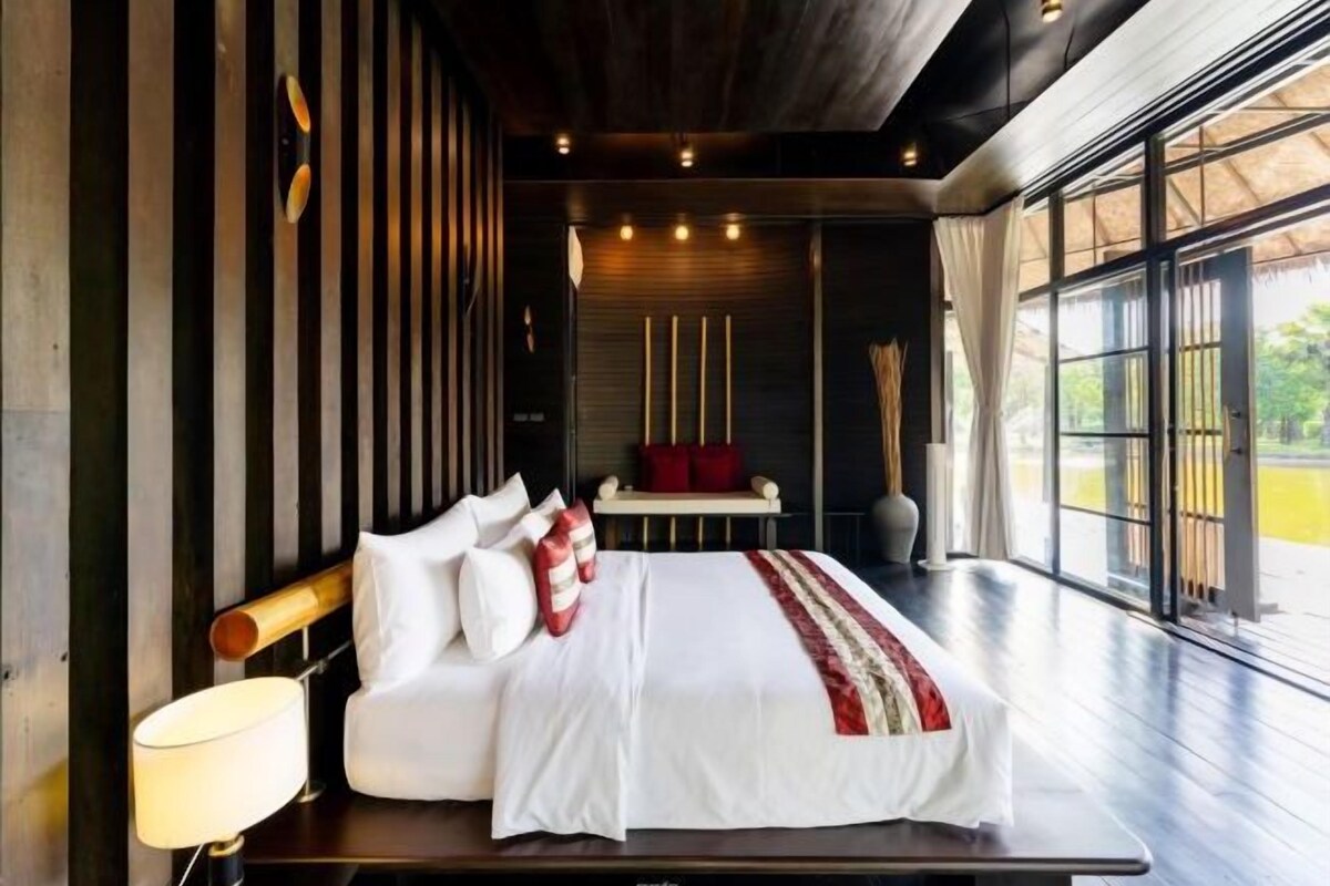 A modern bedroom with a large bed, a bedside table, and a view of the outdoors through large windows.