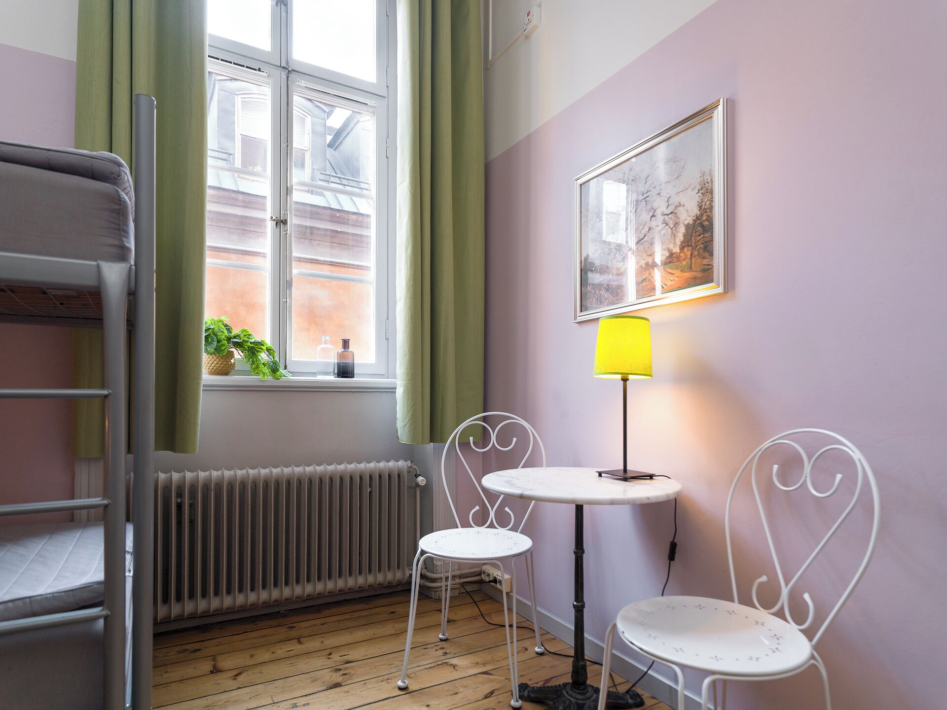 A room with a bunk bed, a radiator, a window with curtains, two white chairs, a small table, and a lamp.