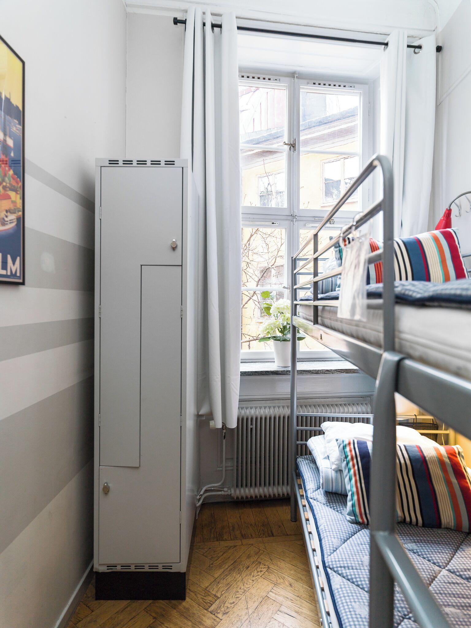 A dormitory room with bunk beds, a radiator, a window with curtains, and a door.