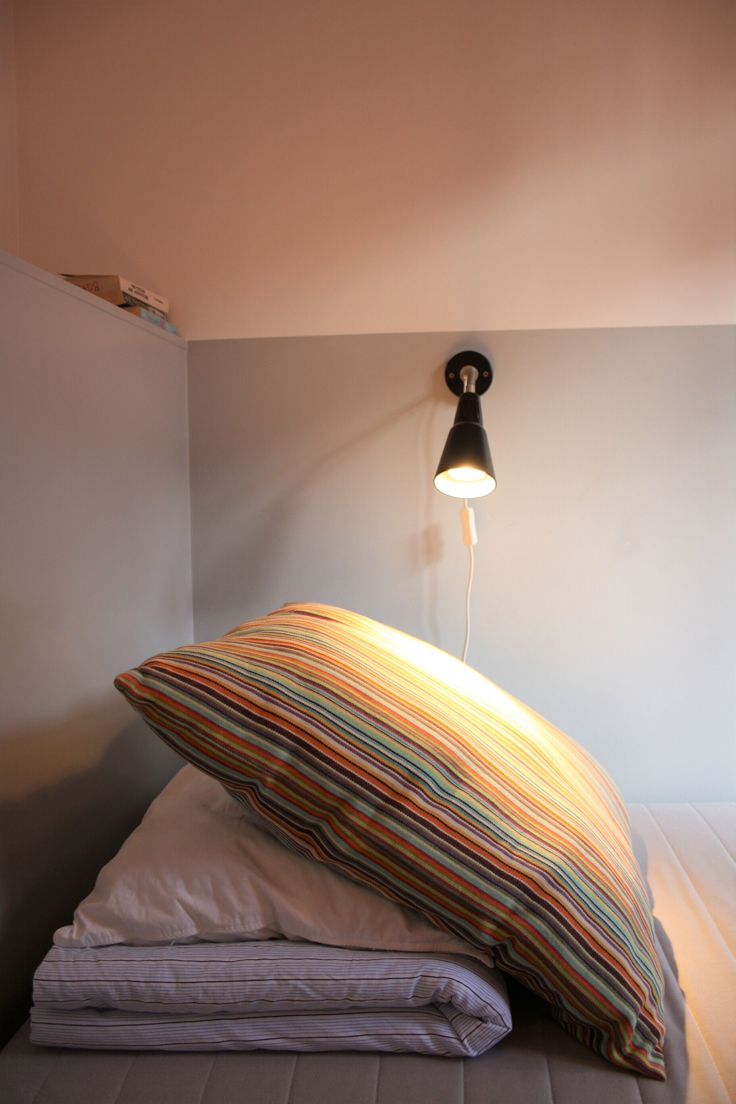 Una cama con un almohadón a rayas y una almohada blanca, una lámpara en la pared y un enchufe.