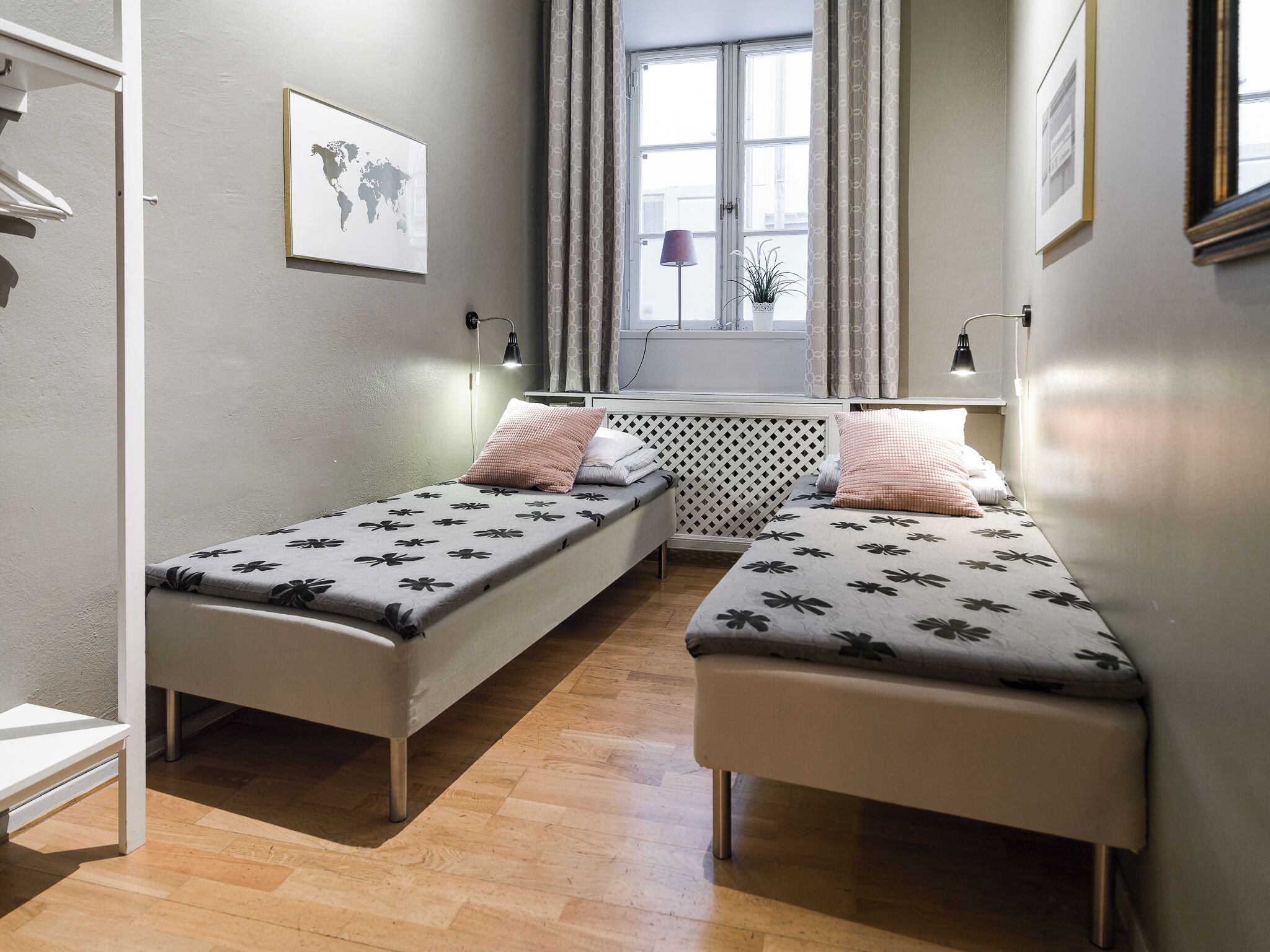 A small, modern room with two single beds, a window with curtains, a world map on the wall, and a radiator.