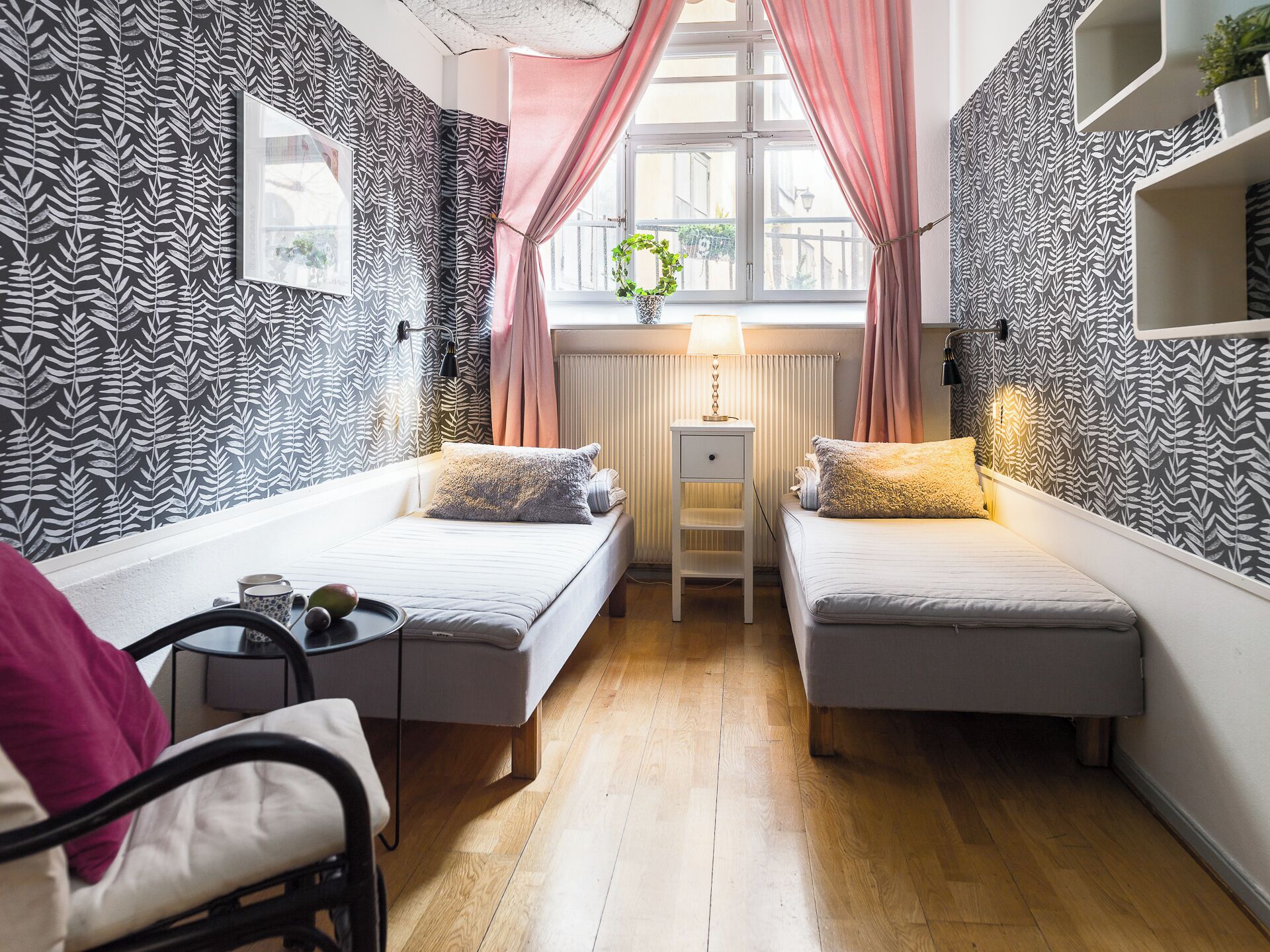 Una habitación pequeña y moderna con dos camas individuales, una silla, una mesa y una ventana con cortinas.