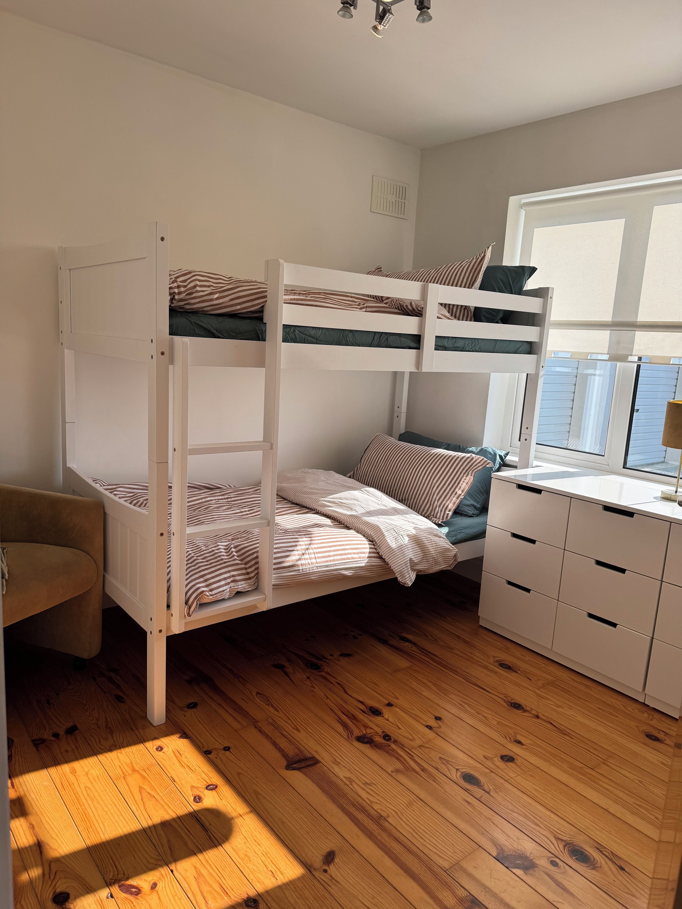 A bunk bed with striped bedding, a wooden desk with drawers, a chair, and a window with blinds.
