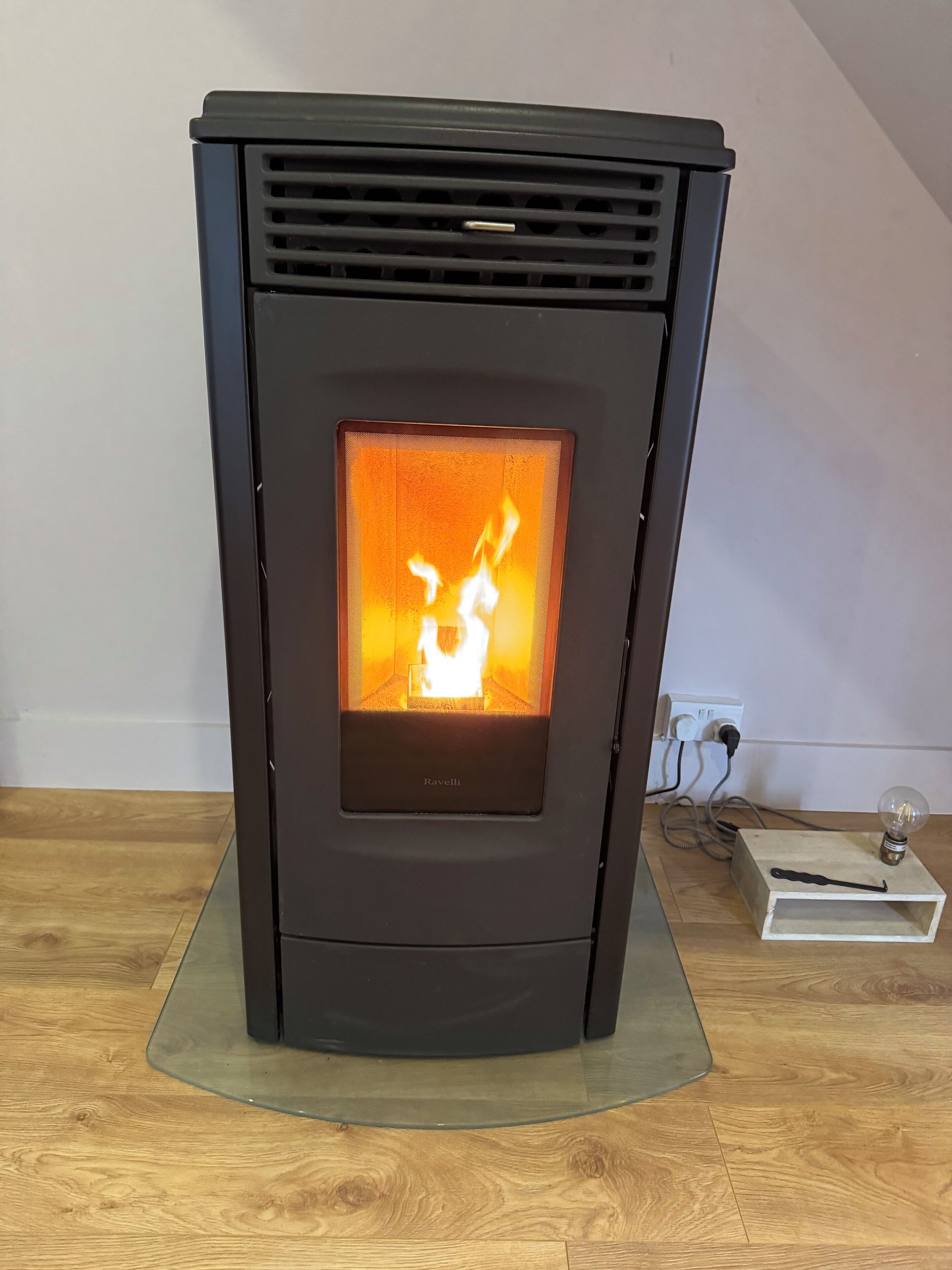 A black wood-burning stove with a glass front, a power outlet, and a light bulb on a wooden floor.