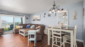 Ein Wohnzimmer mit einem braunen Ecksofa, einem Holztisch, einem Esstisch mit weißen Stühlen und einem großen Fenster mit Blick auf den Strand.