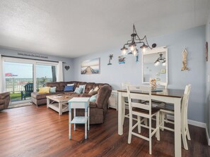 A living room with a brown sectional sofa, a wooden coffee table, a dining table with white chairs, and a large window with a view of the beach. - Starboard Light 7 by Distinctive Beach Rentals (Ormond Beach)