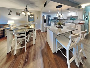 A modern kitchen with white cabinetry, a central island with bar stools, and a dining area with a wooden table and chairs. - Starboard Light 7 by Distinctive Beach Rentals (Ormond Beach)