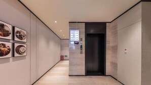 Een moderne hal met een houten vloer, een in de muur gemonteerde lift en ingelijste foto's aan de linkerwand.