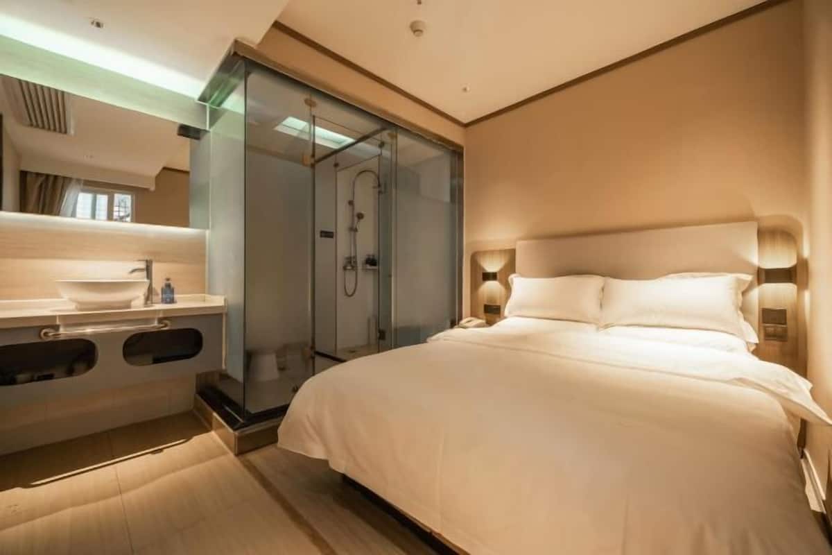A modern hotel room with a large bed, a glass-enclosed shower, and a vanity with a sink and mirror.