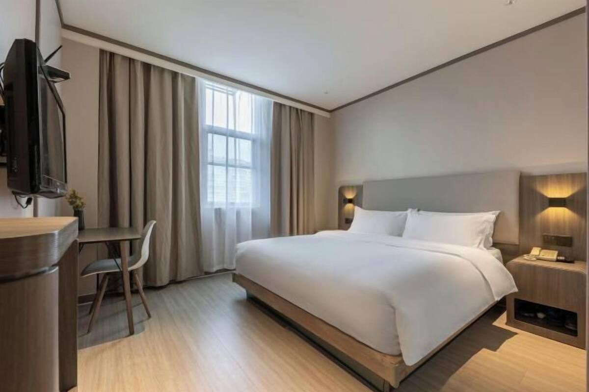 A hotel room with a bed, a desk, a chair, a TV, and a bedside table with a lamp.