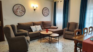 Een woonkamer met een bruine bank, twee fauteuils, een salontafel en een eettafel met stoelen. Twee wandlampen en twee florale schilderijen in lijsten.