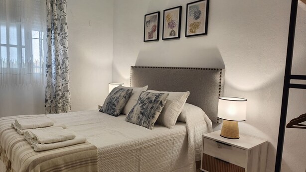 Een net opgemaakt bed met bloemenkussens, een bedlampje en een lijst met een kunstwerk erboven.