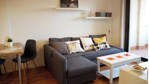 Een moderne woonkamer met een grijze bank, een witte salontafel en een kleine eettafel met stoelen. Aan de muur hangt een schilderij en een raam zorgt voor natuurlijk licht.