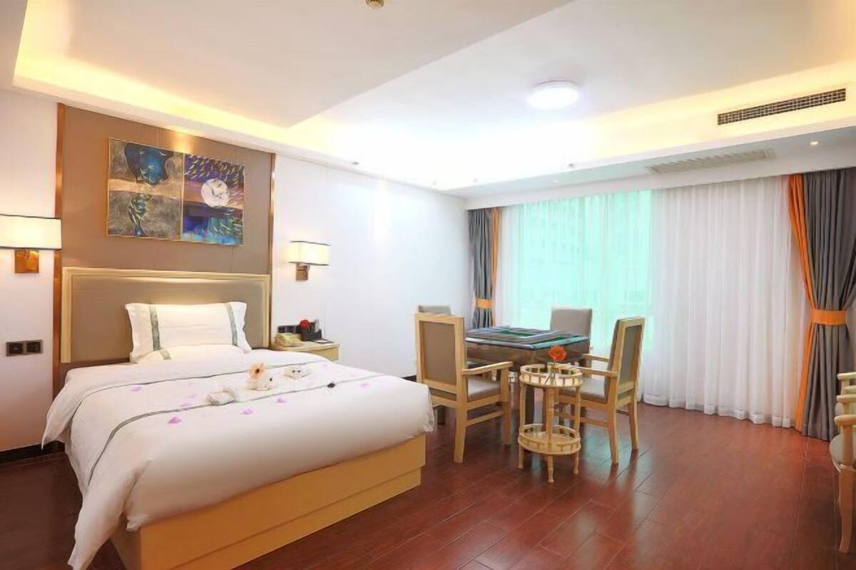 A hotel room with a bed, a desk, chairs, a painting on the wall, and a window with curtains.