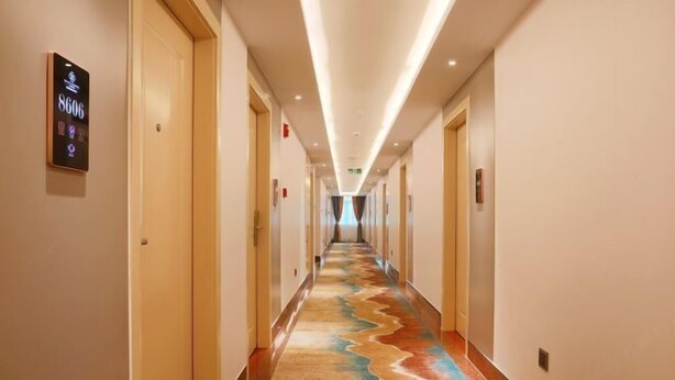 Un couloir d’hôtel avec un panneau numérique indiquant le numéro des chambres, des portes en bois et une moquette colorée.