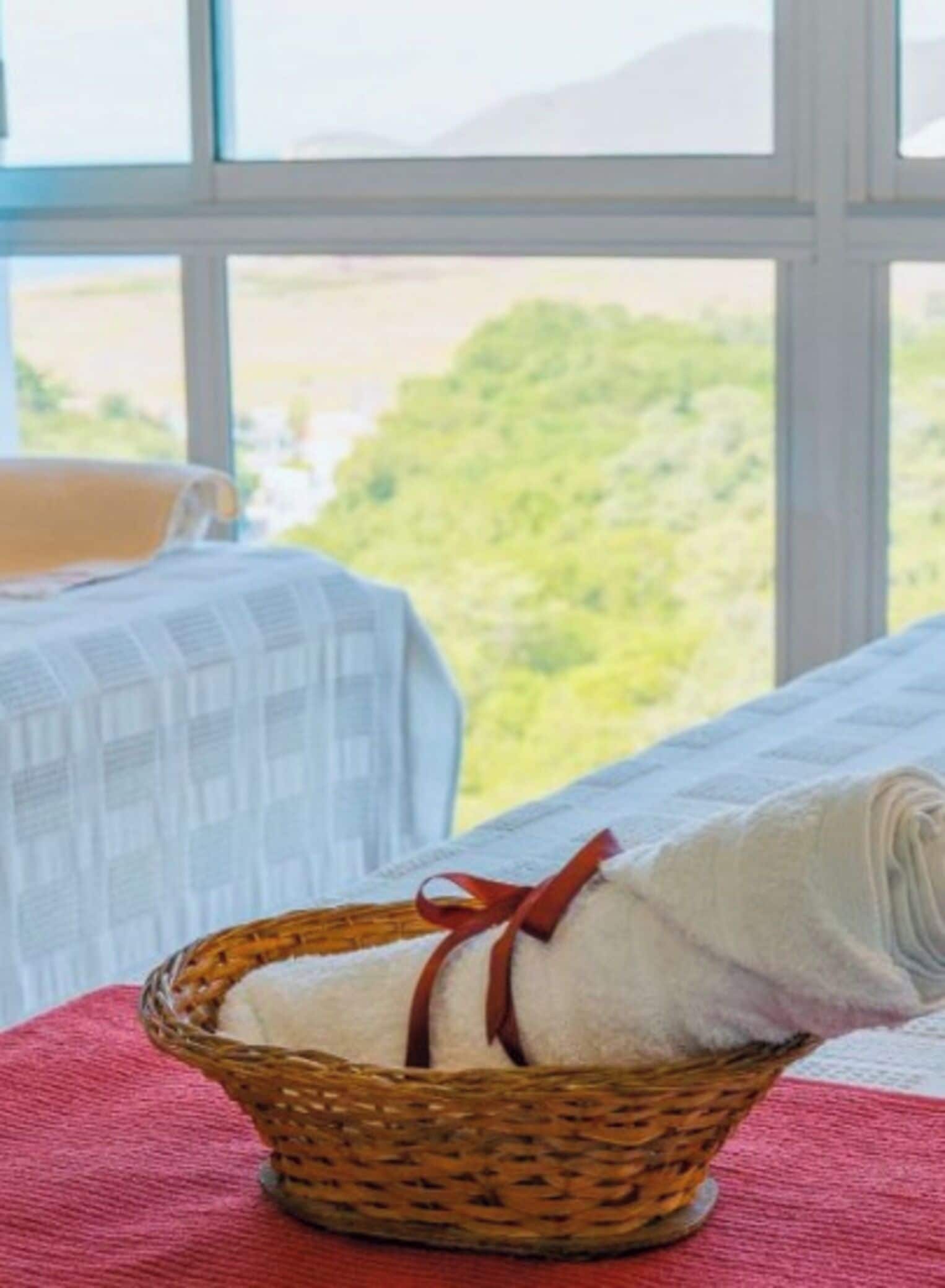 A room with a large window, a wicker basket with a towel, and a bed with a red blanket.