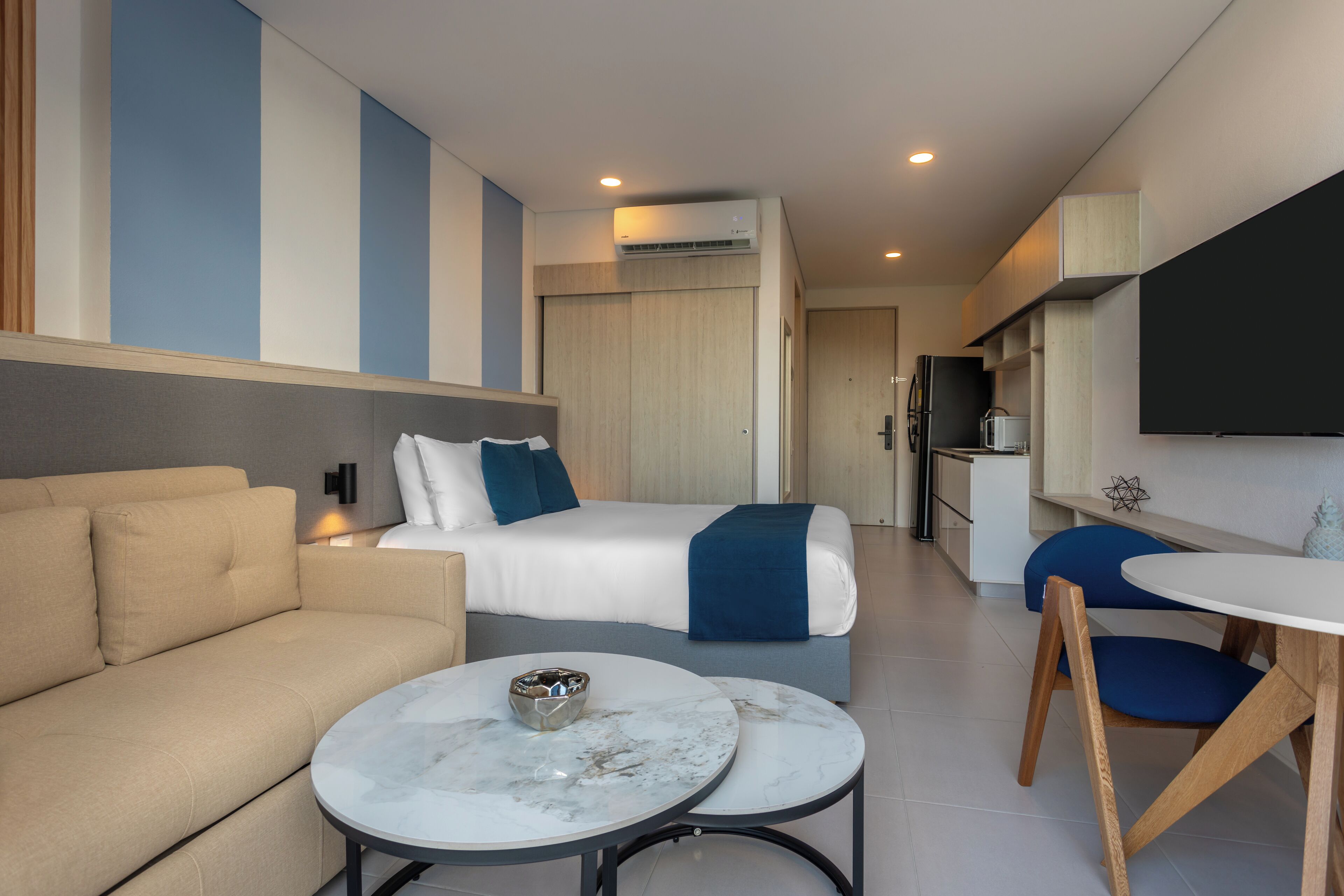 Habitació d'hotel moderna amb un sofà, un llit amb una manta blava, una taula de dinar i una televisió.